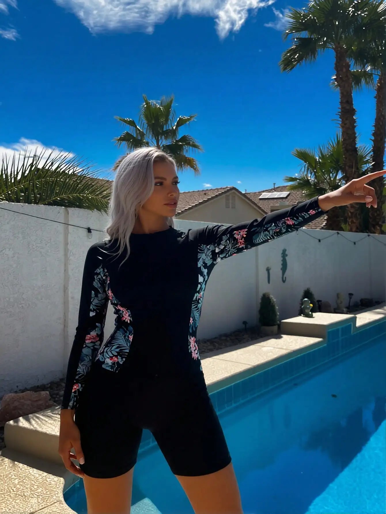 long sleeve floral rashguard swimsuit worn by a woman pointing near a swimming pool on a sunny day.